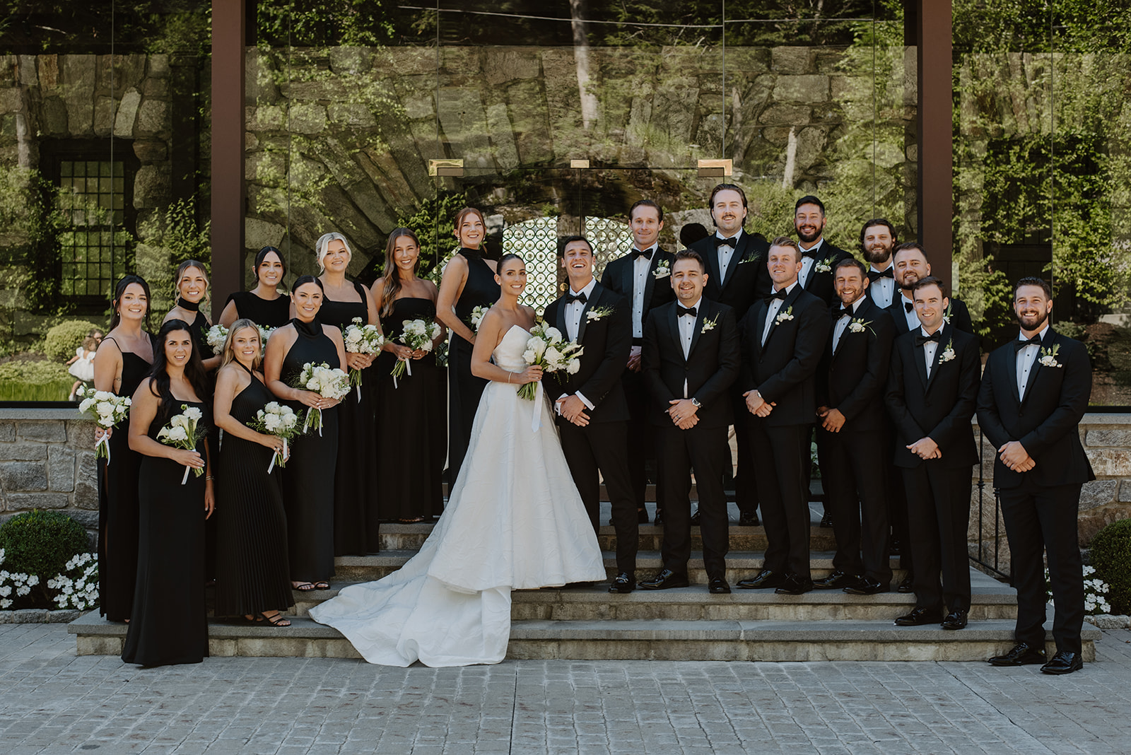 Full wedding party posing on stone steps, bridesmaids in black dresses and groomsmen in classic tuxedos.